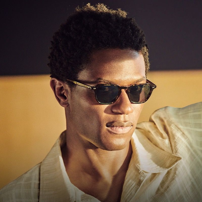 Close-up studio shot of an African-American man wearing sunglasses and a tan shirt against a split black and tan background.