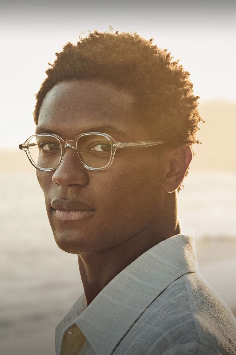 Close-up of a dark-skinned man with clear eyeglasses and a light blue shirt standing outside, bathed in warm sunlight.