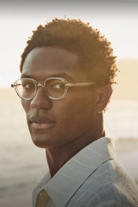Portrait of Hamid Onifadé wearing clear-framed glasses and a light collared shirt against a bright sunset backdrop by Matthew Brookes.