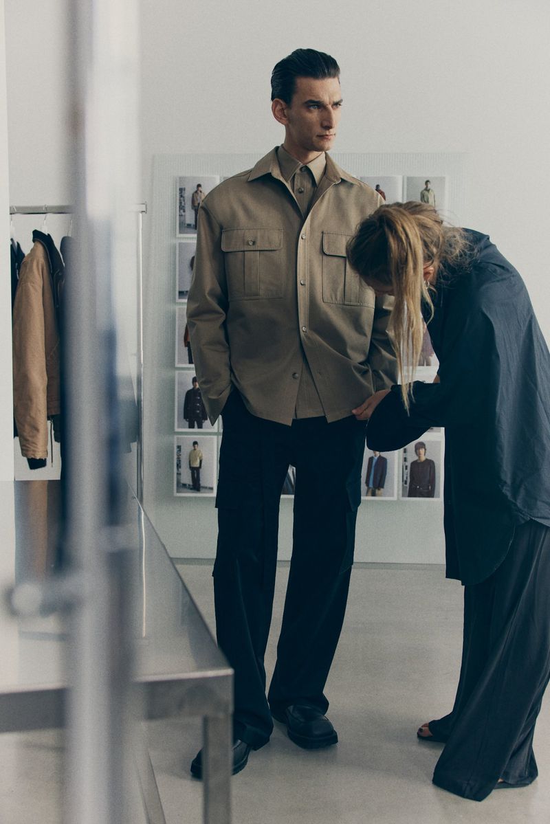 A male fashion model poses indoors while a stylist adjusts his shirt; he wears a tan shirt, dark trousers, and faces the left.