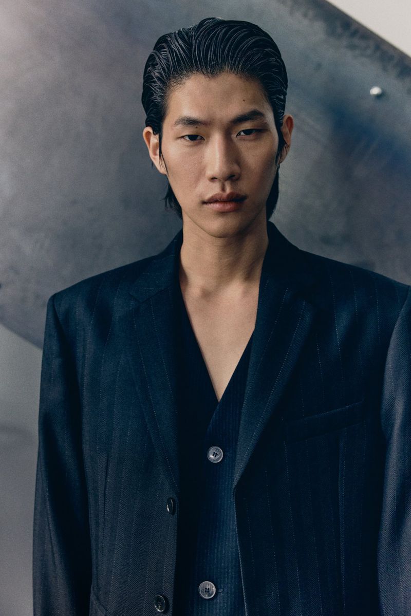 Portrait of an Asian male model wearing a dark suit with a vest against a grey background in a studio with soft, diffused lighting.