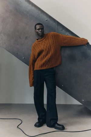 A black male model is leaning against a metal staircase wearing a brown cable knit sweater, black pants, and black shoes in a studio.