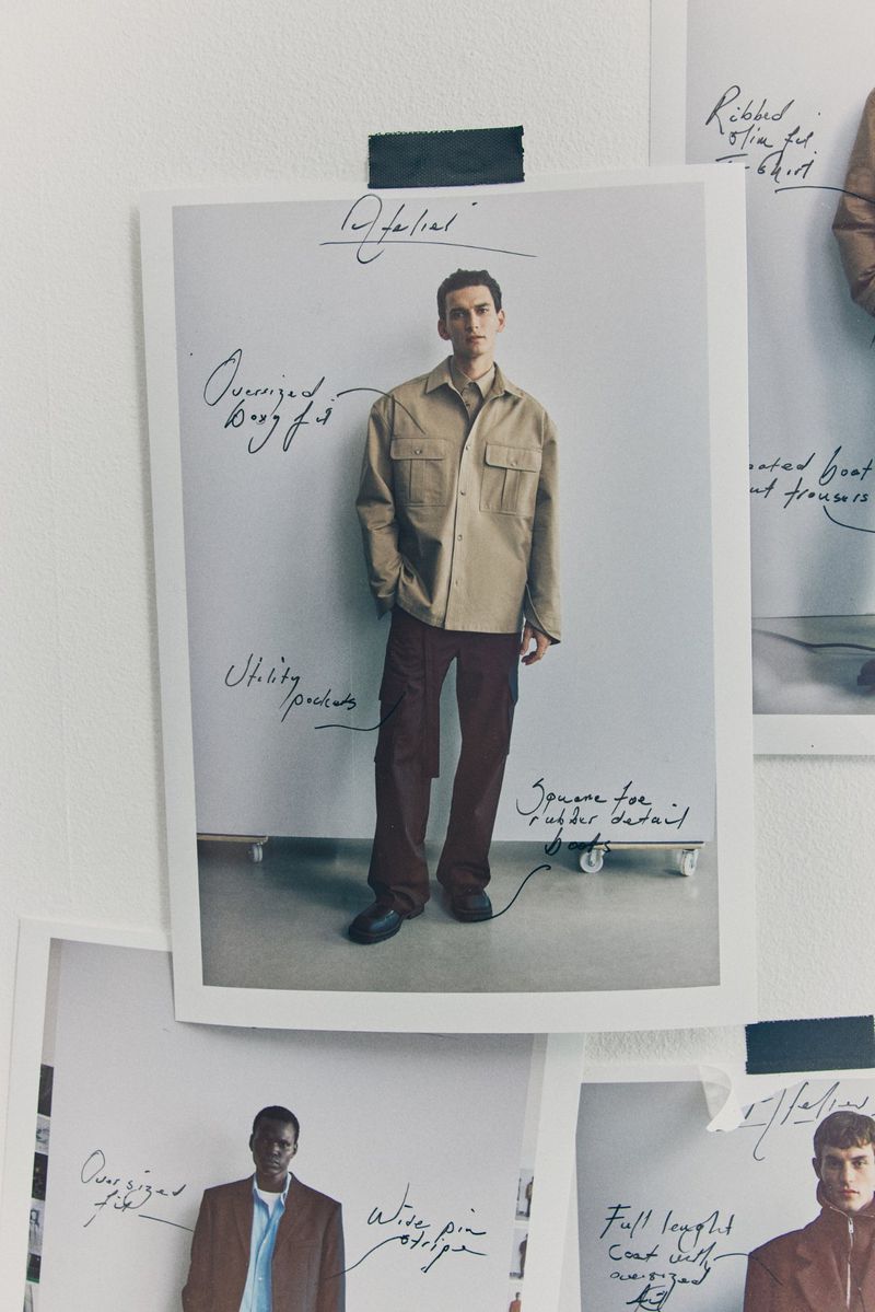 Photo pinned to a white wall shows a male model wearing a tan shirt jacket and burgundy pants, surrounded by fashion sketches.