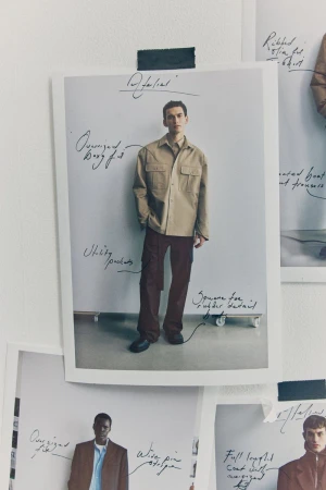 Photo pinned to a white wall shows a male model wearing a tan shirt jacket and burgundy pants, surrounded by fashion sketches.