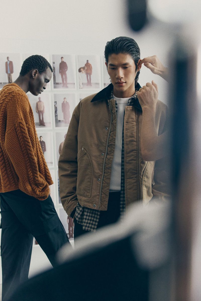Two men model tailored, casual fashion in a neutral indoor studio. One wears a brown cable-knit sweater, and the other wears a brown jacket.
