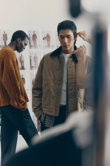 Two men model tailored, casual fashion in a neutral indoor studio. One wears a brown cable-knit sweater, and the other wears a brown jacket.