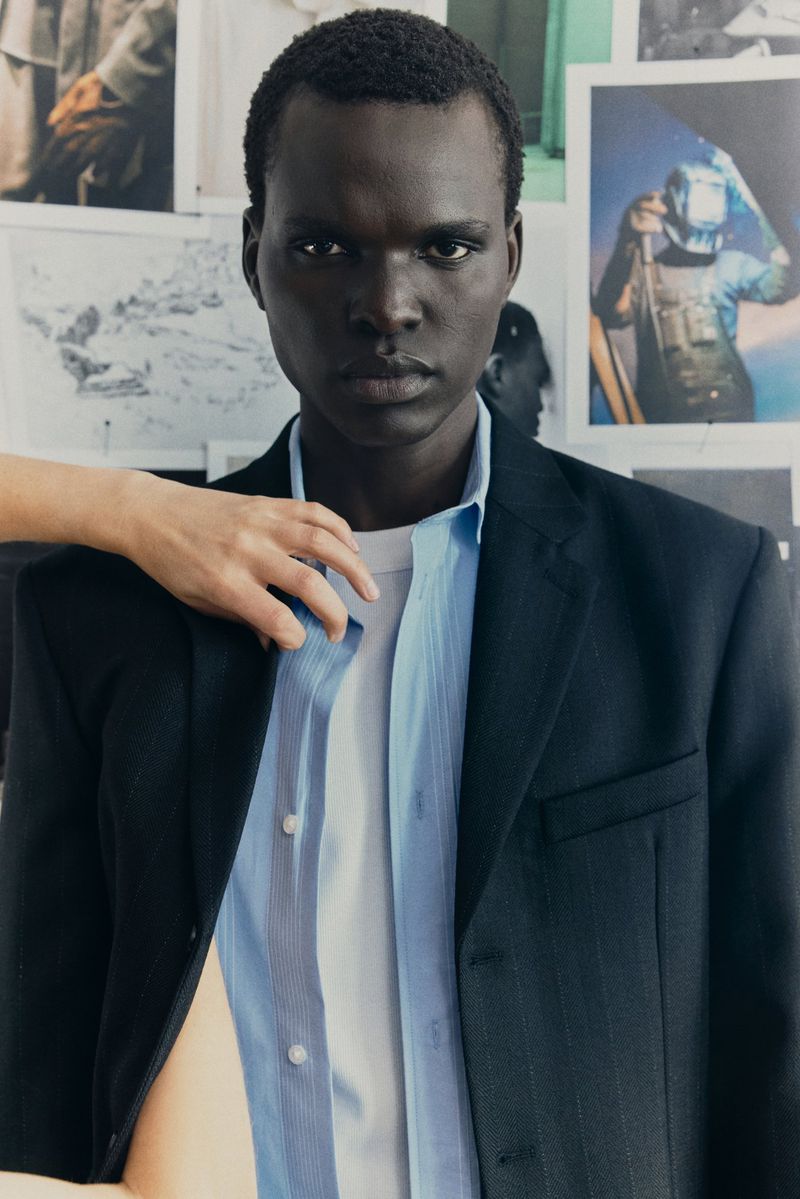 Close-up of a dark-skinned man in an open blazer with a hand on his shoulder against a background of tacked-up photos.