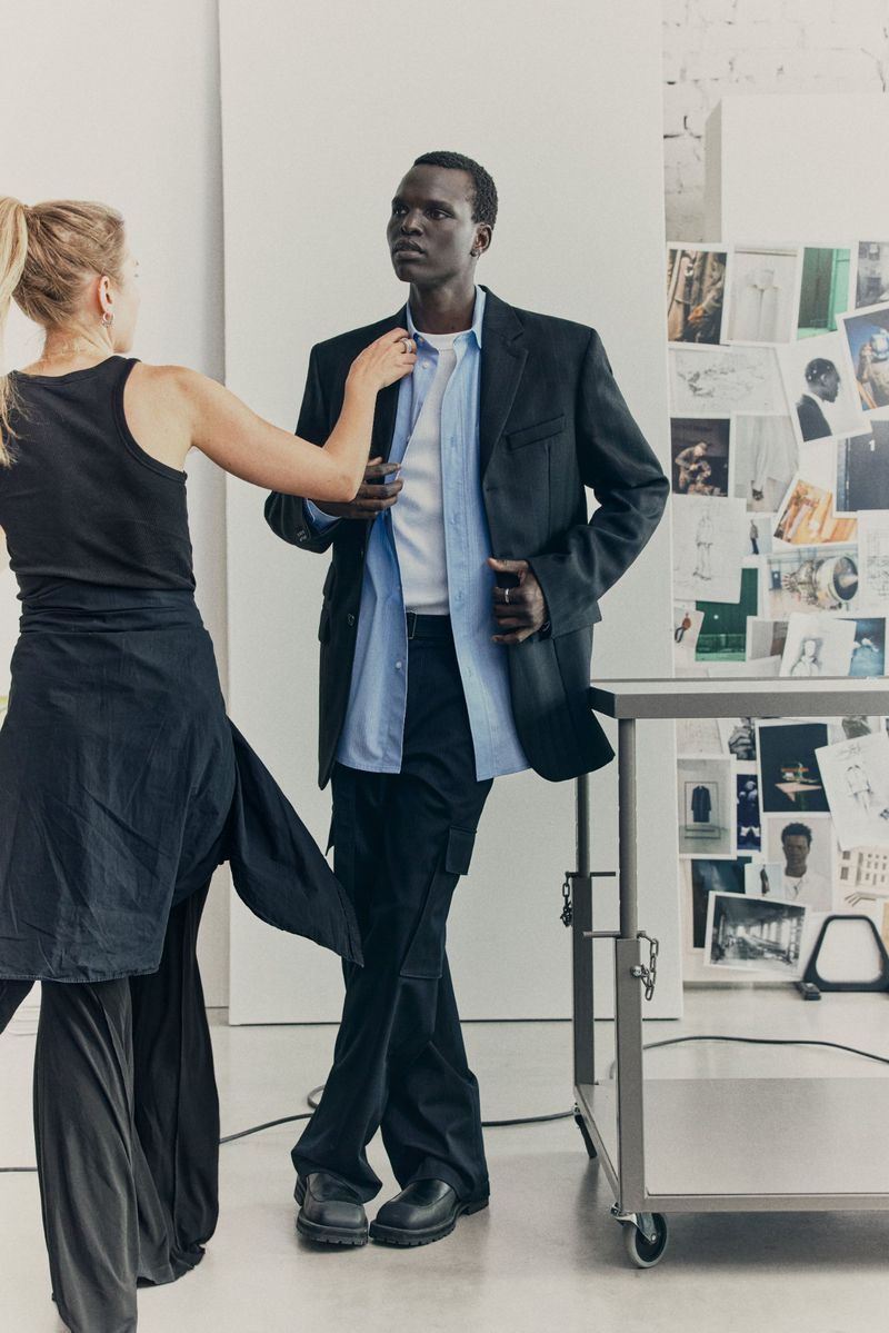 Dark-skinned male fashion model in blazer and dress pants adjusts his light-blue button-up shirt in a photo studio with assistant.