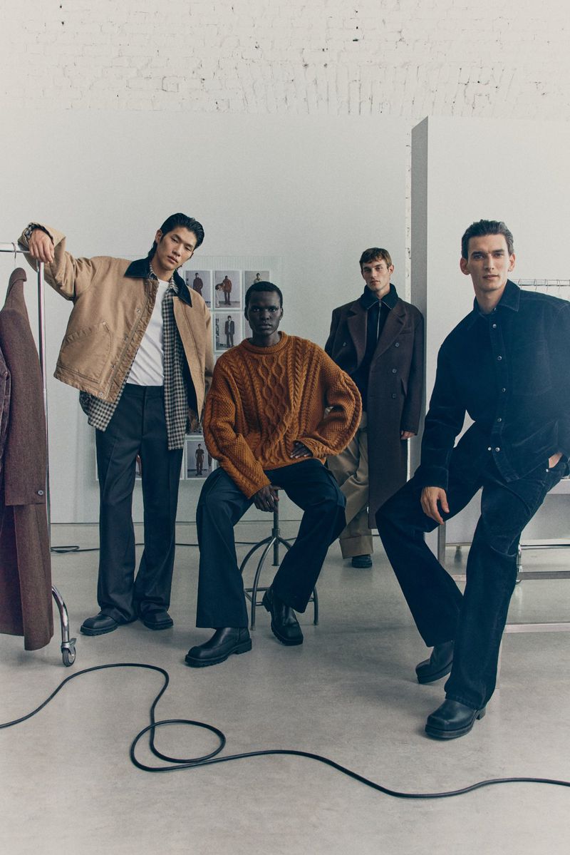Four male fashion models in an indoor studio setting wearing modern jackets, sweaters, and trousers in a fashion editorial shot.
