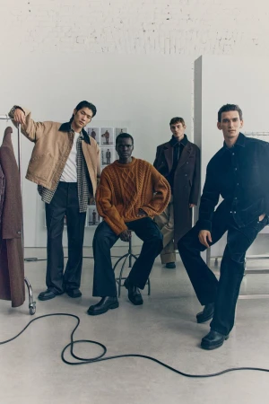 Four male fashion models in an indoor studio setting wearing modern jackets, sweaters, and trousers in a fashion editorial shot.