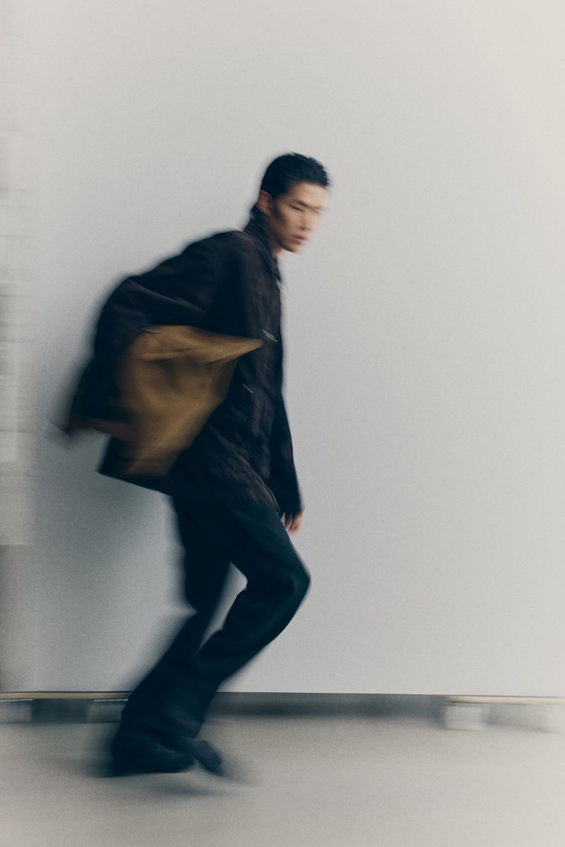 A blurred, medium shot of a stylish adult male in a dark jacket and pants, walking against a white backdrop in a fashion photography setting.