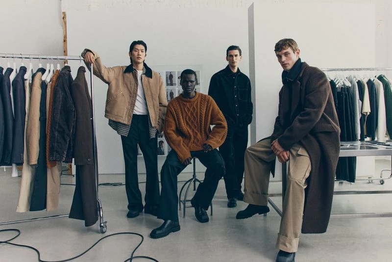 Group of four adult male fashion models posing indoors, wearing sweaters, jackets, coats, and trousers, near clothing rack and metal table.