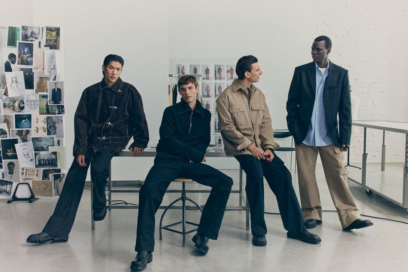 Four adult male fashion models sit and stand in a studio wearing dark and neutral-toned jackets and trousers in a bright and airy setting.