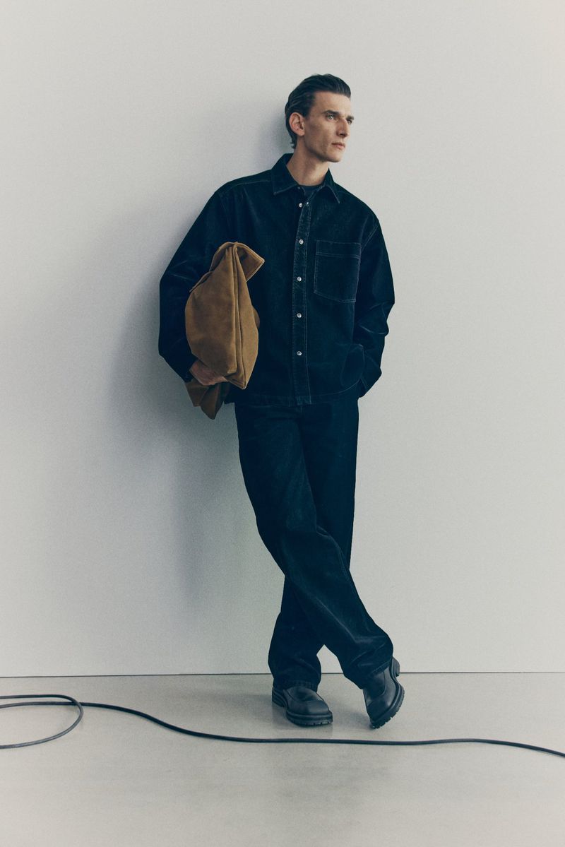 Stylish adult man in a relaxed pose indoors, wearing a dark button-down shirt and jeans, holding a tan suede bag against a plain background.