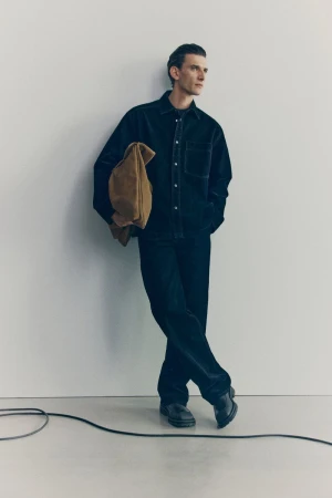 Stylish adult man in a relaxed pose indoors, wearing a dark button-down shirt and jeans, holding a tan suede bag against a plain background.