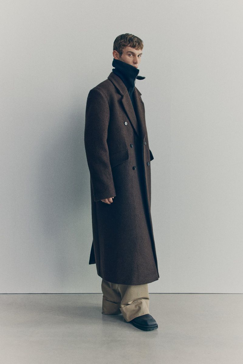 Man in brown overcoat and turtleneck sweater poses in a studio. He wears beige trousers and black shoes against a light-grey backdrop.