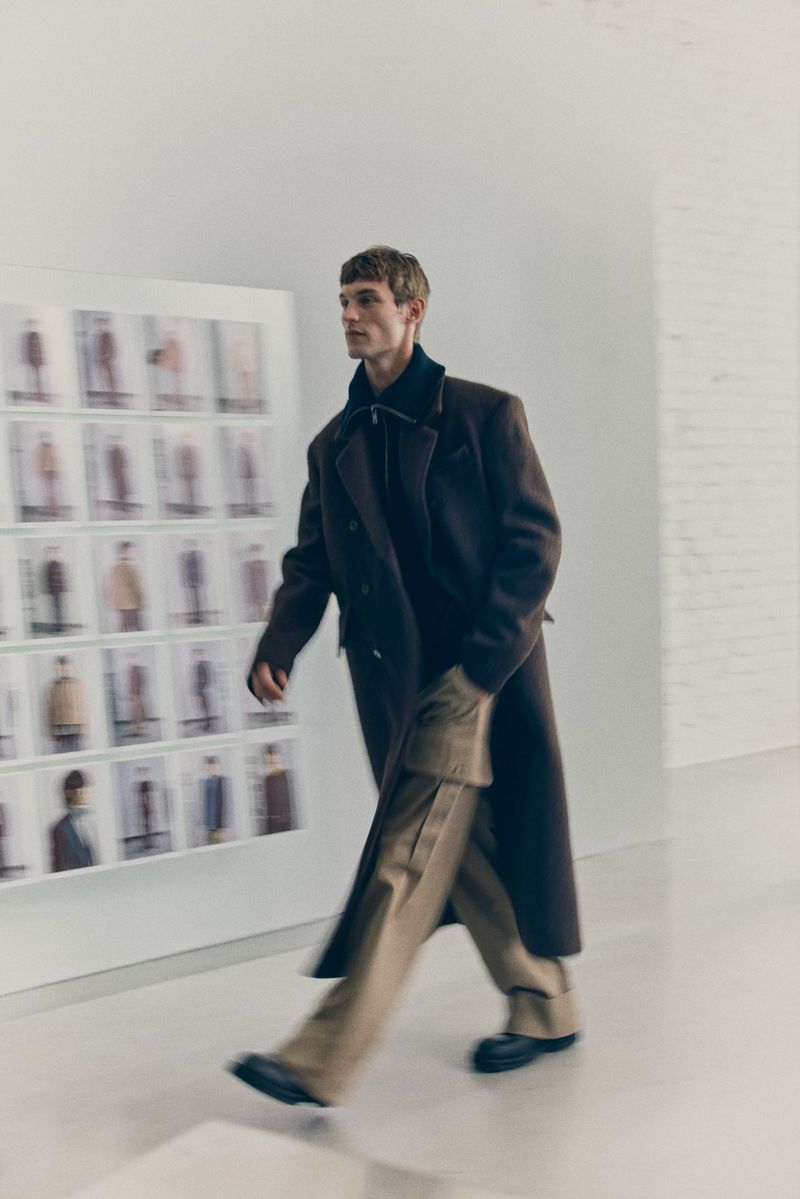 A male fashion model walks in a runway show wearing a long brown wool coat and tan trousers with pockets, posing against a wall.