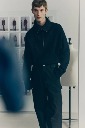 A male fashion model is posing indoors wearing a ribbed, navy-blue, quarter-zip sweater and matching black pants against a bright background.