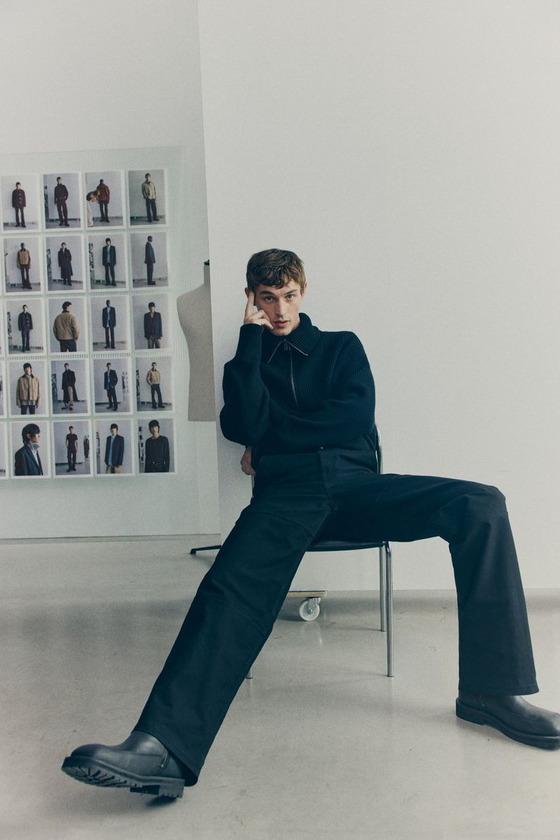 Male model seated on a metal chair wearing a dark sweater and trousers against a white wall with photo grid