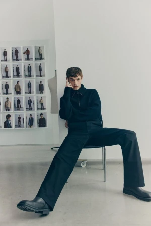 Male model seated on a metal chair wearing a dark sweater and trousers against a white wall with photo grid