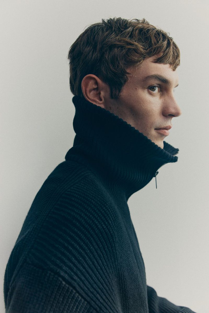 Profile portrait of a young adult man with brown hair wearing a dark blue ribbed knit turtleneck sweater against a neutral background.