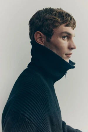 Profile portrait of a young adult man with brown hair wearing a dark blue ribbed knit turtleneck sweater against a neutral background.
