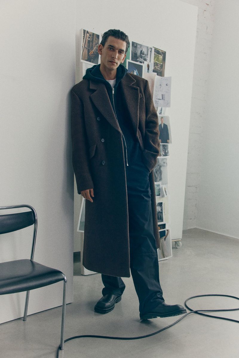 A young man stands in a long brown coat against a white wall with pinned images, styled with a hoodie, dark pants, and black leather shoes.