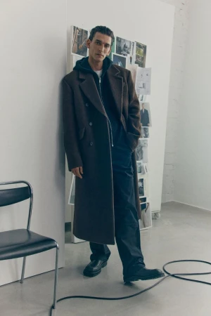 A young man stands in a long brown coat against a white wall with pinned images, styled with a hoodie, dark pants, and black leather shoes.