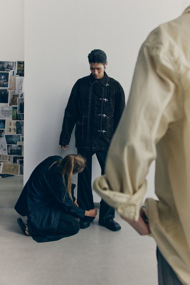 A casually dressed man stands while a woman adjusts his pants hem; another person’s arm is blurred in the foreground in a bright indoor setting.