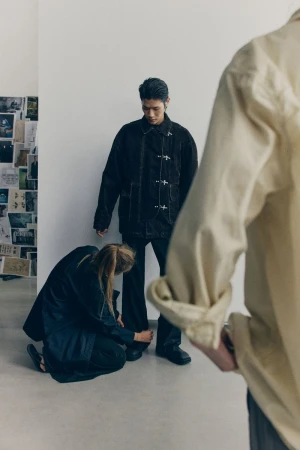 A casually dressed man stands while a woman adjusts his pants hem; another person’s arm is blurred in the foreground in a bright indoor setting.