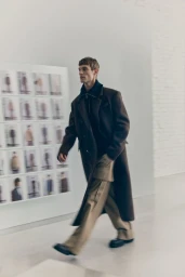 Kit Butler walking in a long brown coat and wide-leg beige trousers past a moodboard wall for H&M Atelier A/W 2025.