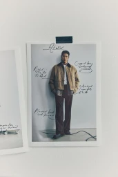 Fashion photograph of a male model wearing a tan jacket and dark trousers pinned to a plain white wall