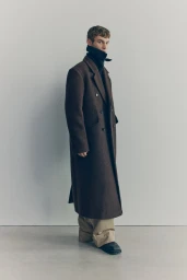 Model Kit Butler in a long brown wool overcoat and wide-leg beige trousers in a minimalist studio for H&M Atelier A/W 2025.