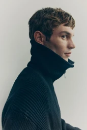 Side profile portrait of model Kit Butler wearing a dark ribbed high-neck zip sweater against a pale background.