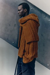 Model Badhiel Lony Nyang wears a textured caramel-brown cable-knit sweater and draped hood in profile against an angled dark wall.