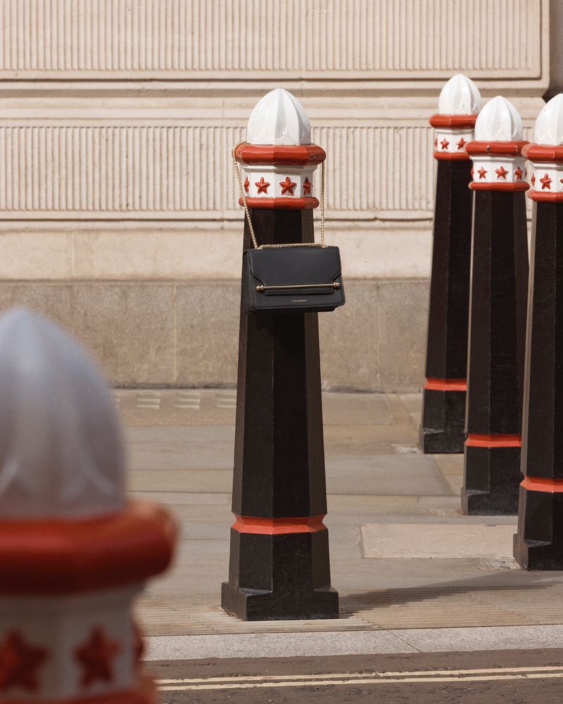 Black purse with gold chain strap hanging from a bollard with red and white details in an urban outdoor setting on a sunny day.