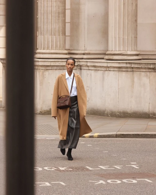 Woman in tan coat walks across city street with building and columns in background; she carries a brown purse over her shoulder.