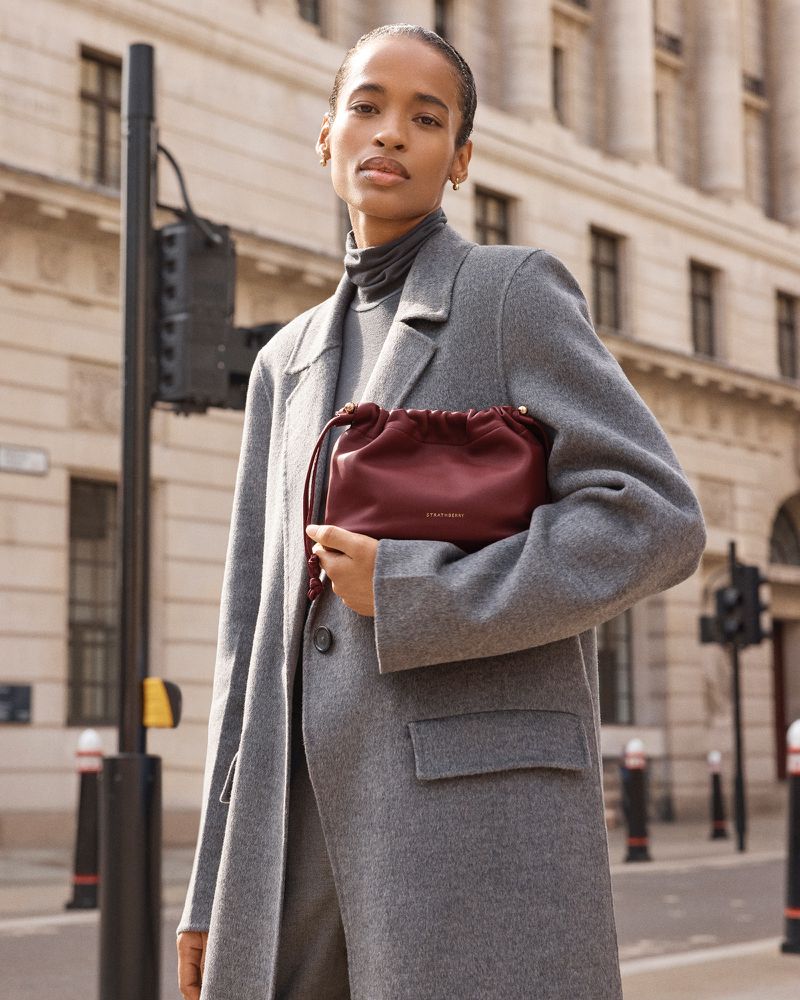 Woman in a grey coat poses on a city street, holding a burgundy drawstring bag. Urban fashion shot with architectural background and traffic signal.