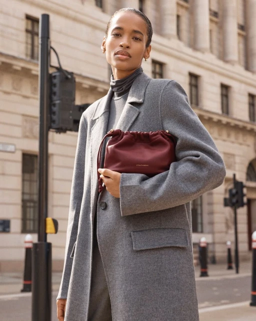 Woman in a grey coat poses on a city street, holding a burgundy drawstring bag. Urban fashion shot with architectural background and traffic signal.