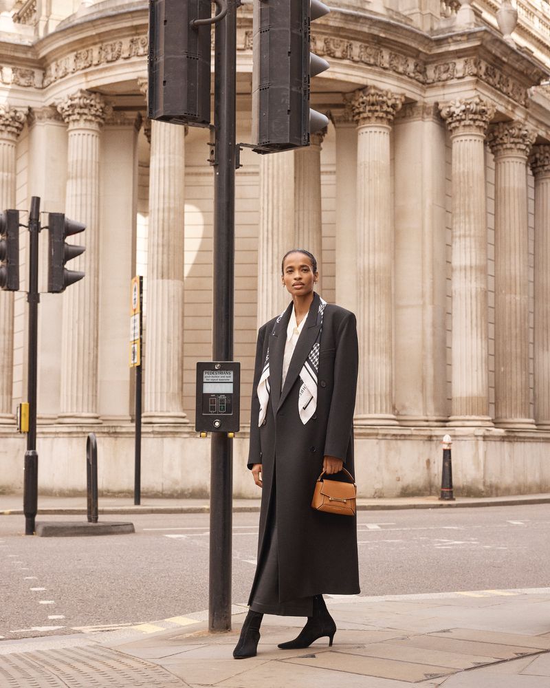 A woman in a coat and scarf stands on a city street near a traffic light and a building with large columns in the background.
