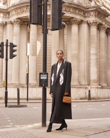 A woman in a coat and scarf stands on a city street near a traffic light and a building with large columns in the background.