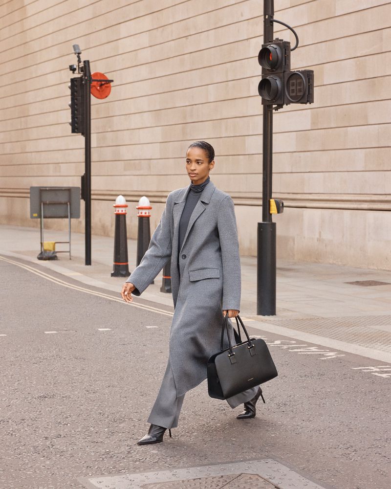 An adult female model in a gray coat and black heels strides across a crosswalk holding a black handbag in a city street.