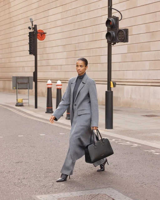 An adult female model in a gray coat and black heels strides across a crosswalk holding a black handbag in a city street.