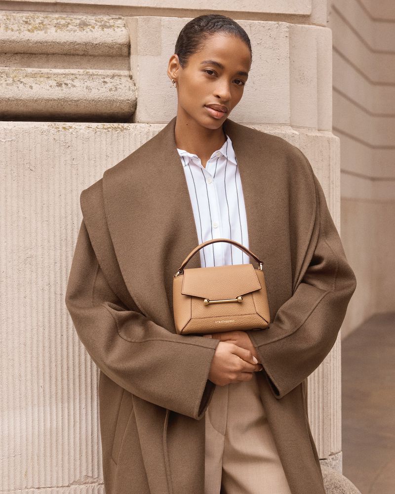 Adult female model in a brown coat holding a tan leather handbag with gold hardware posing against a beige textured wall.