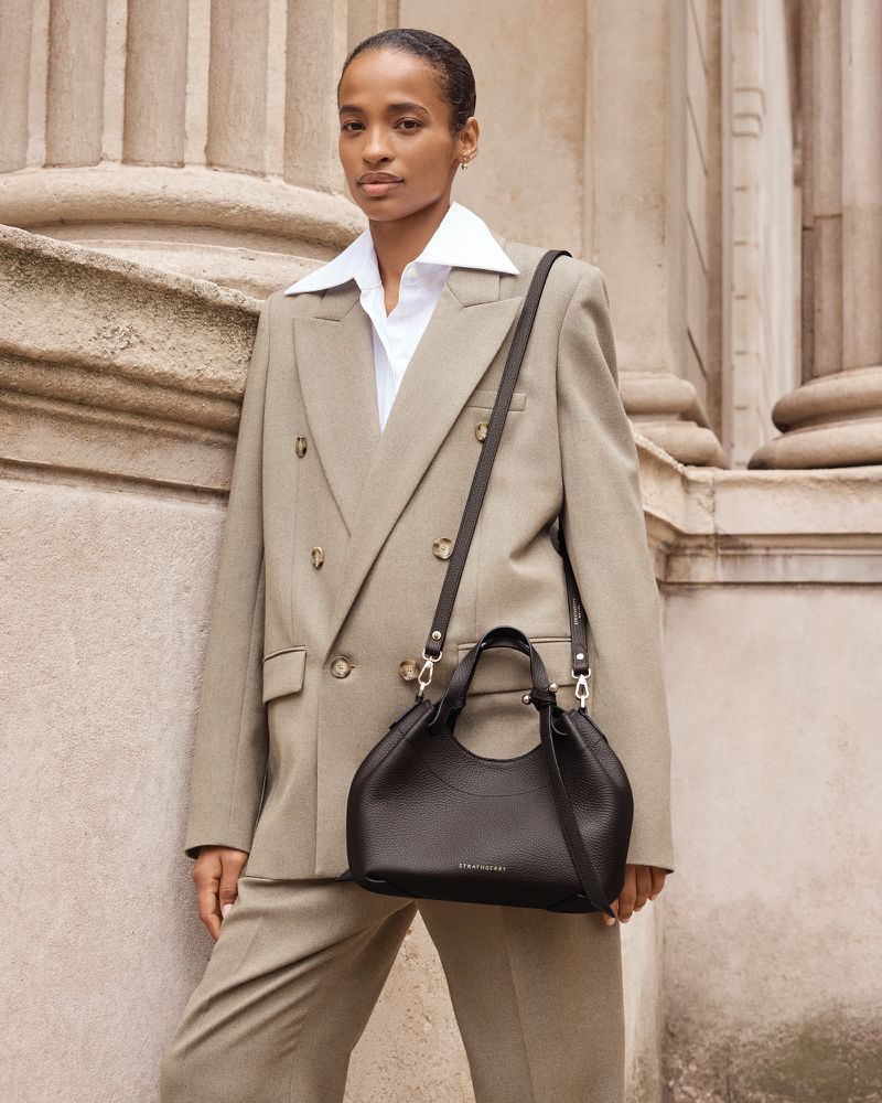 Woman in a tan suit and white button-down shirt carrying a black leather purse in front of a stone facade, neutral tones.