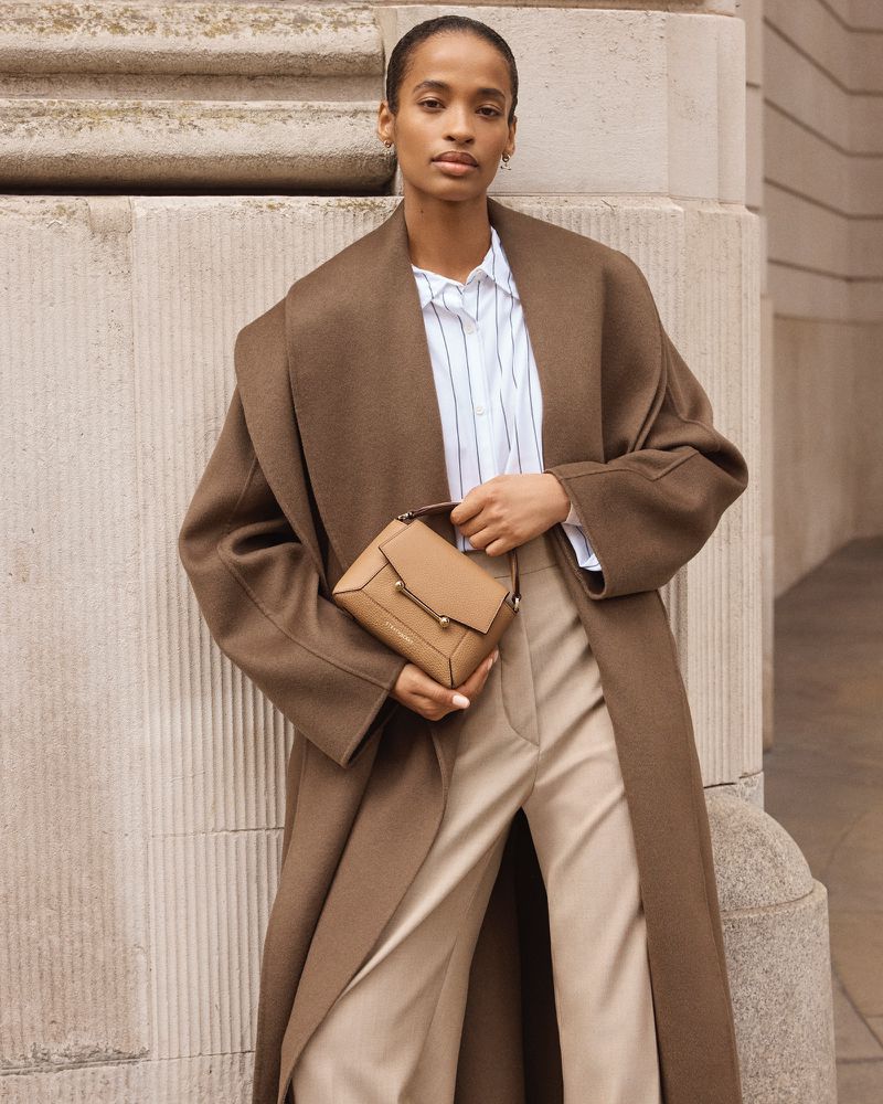 Adult woman wearing a brown trench coat and carrying a tan purse poses near a grey concrete wall in an outdoor setting.