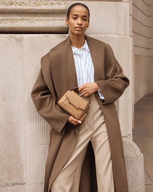 Adult woman wearing a brown trench coat and carrying a tan purse poses near a grey concrete wall in an outdoor setting.