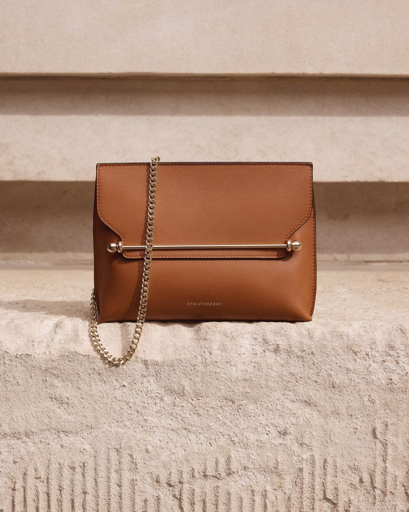 Brown leather Strathberry handbag with gold hardware and chain strap resting on stone steps. Close-up studio product shot for fashion.