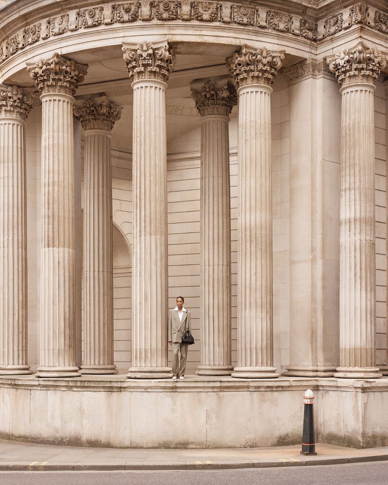 Adult male in suit standing in front of building with stone columns, holding a bag in an outdoor, daytime architectural setting.