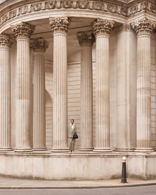 Adult male in suit standing in front of building with stone columns, holding a bag in an outdoor, daytime architectural setting.